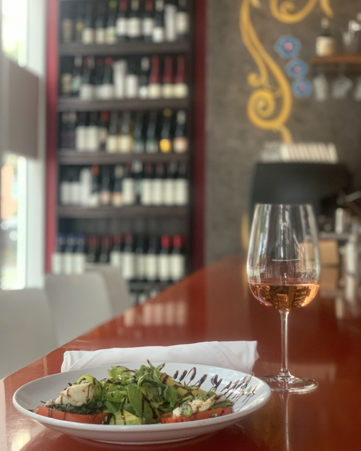 Caprese salad and a glass of rosé on the red bar top, wine wall behind — daytime at Renzo's Wine Bar, St. Petersburg